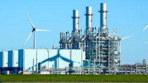 A view of a power plant with wind mills surrounding it.